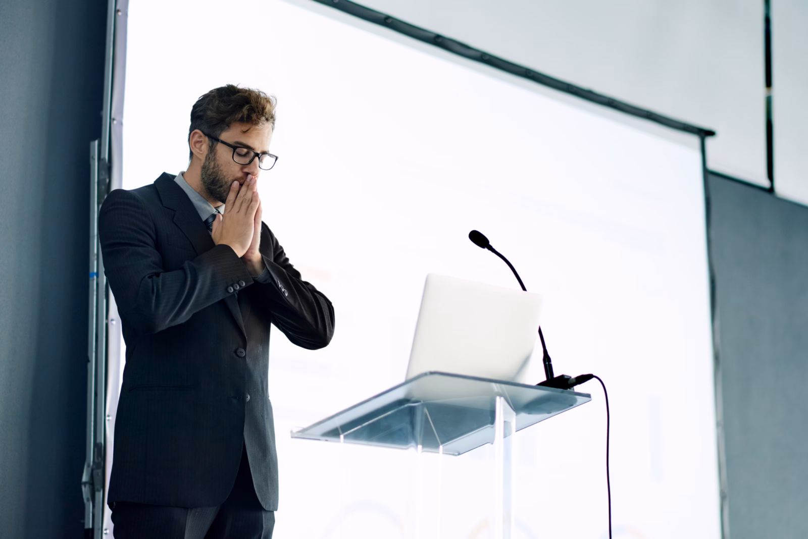 A man in a suit stands in front of a lectern with his hands over his mouth and his eyes closed.