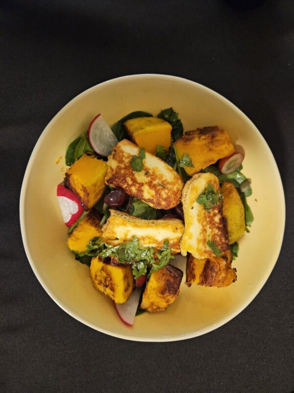 Round yellow plate holding a salad of cooked pumpkin, radishes, halloumi and greens.