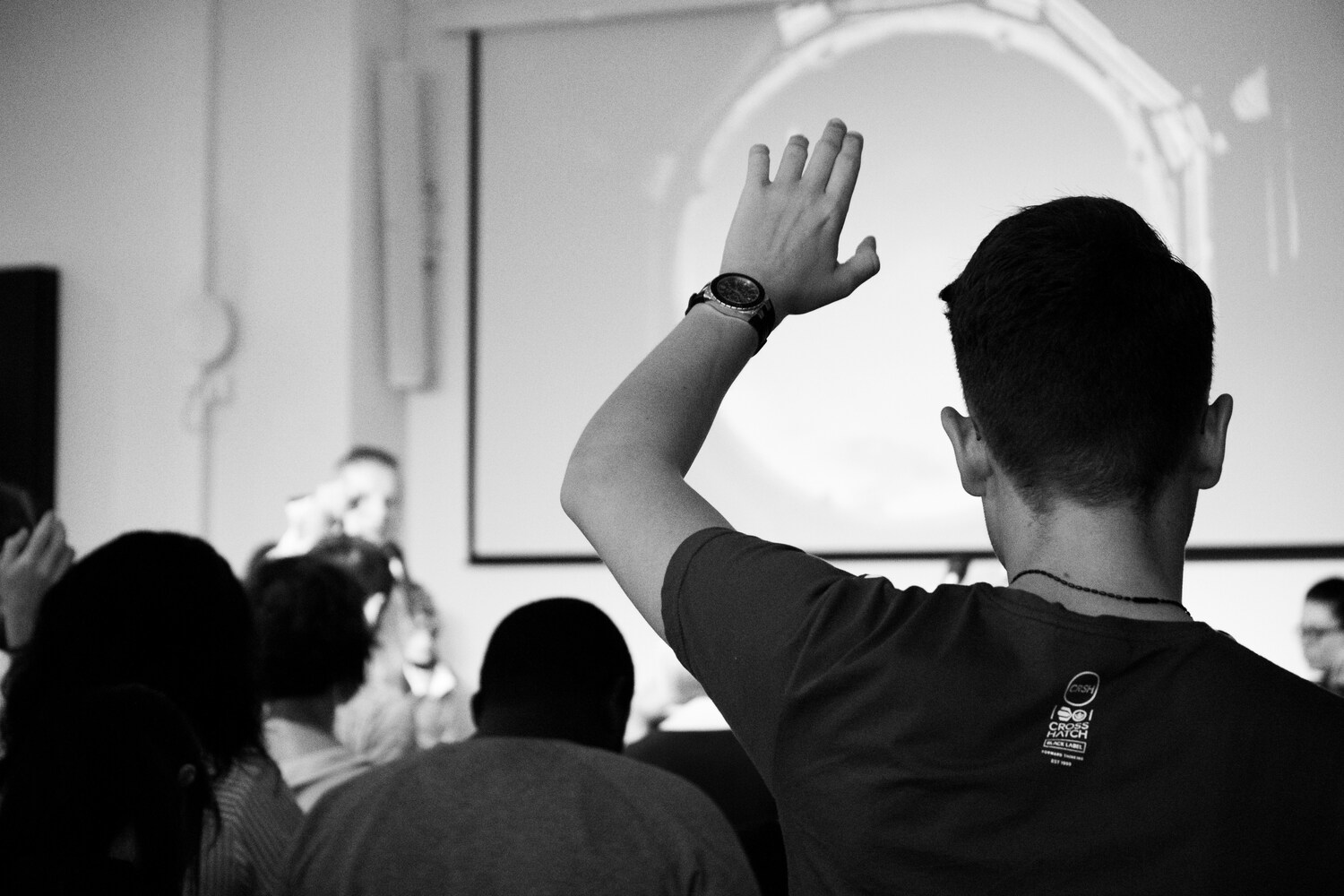 A man in an audience seen from behind, raising his hand to a presenter on stage.