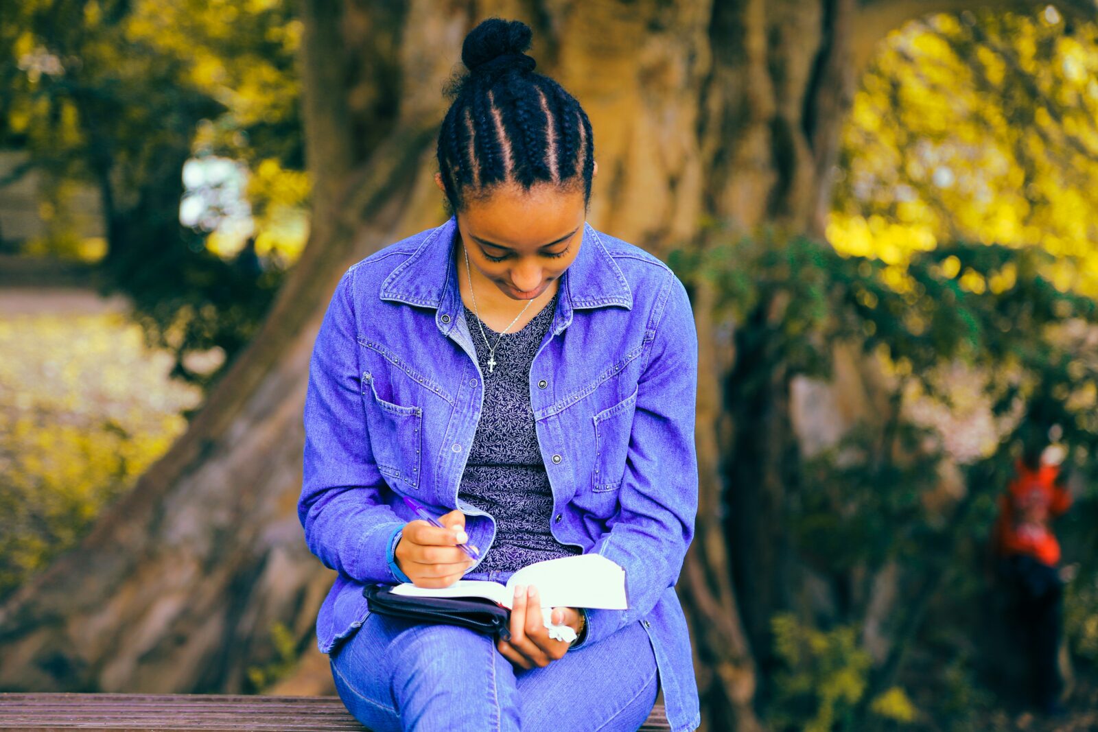 A young woman dressed in denim sits in front of a tree writing in a book