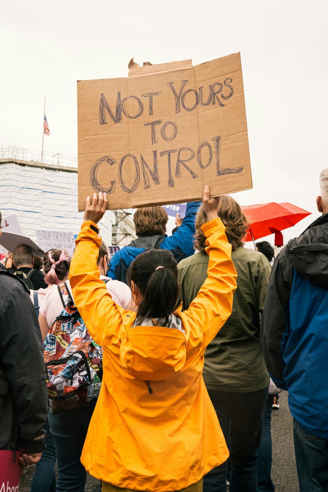 A woman in a yellow jacket, seen from the back, holds a sign reading, “Not Yours to Control”