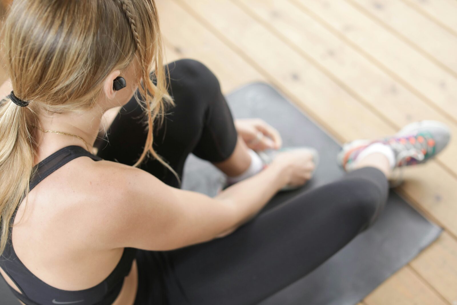 A woman in exercise gear, seen from above, sits on a yoga mat and ties one shoe