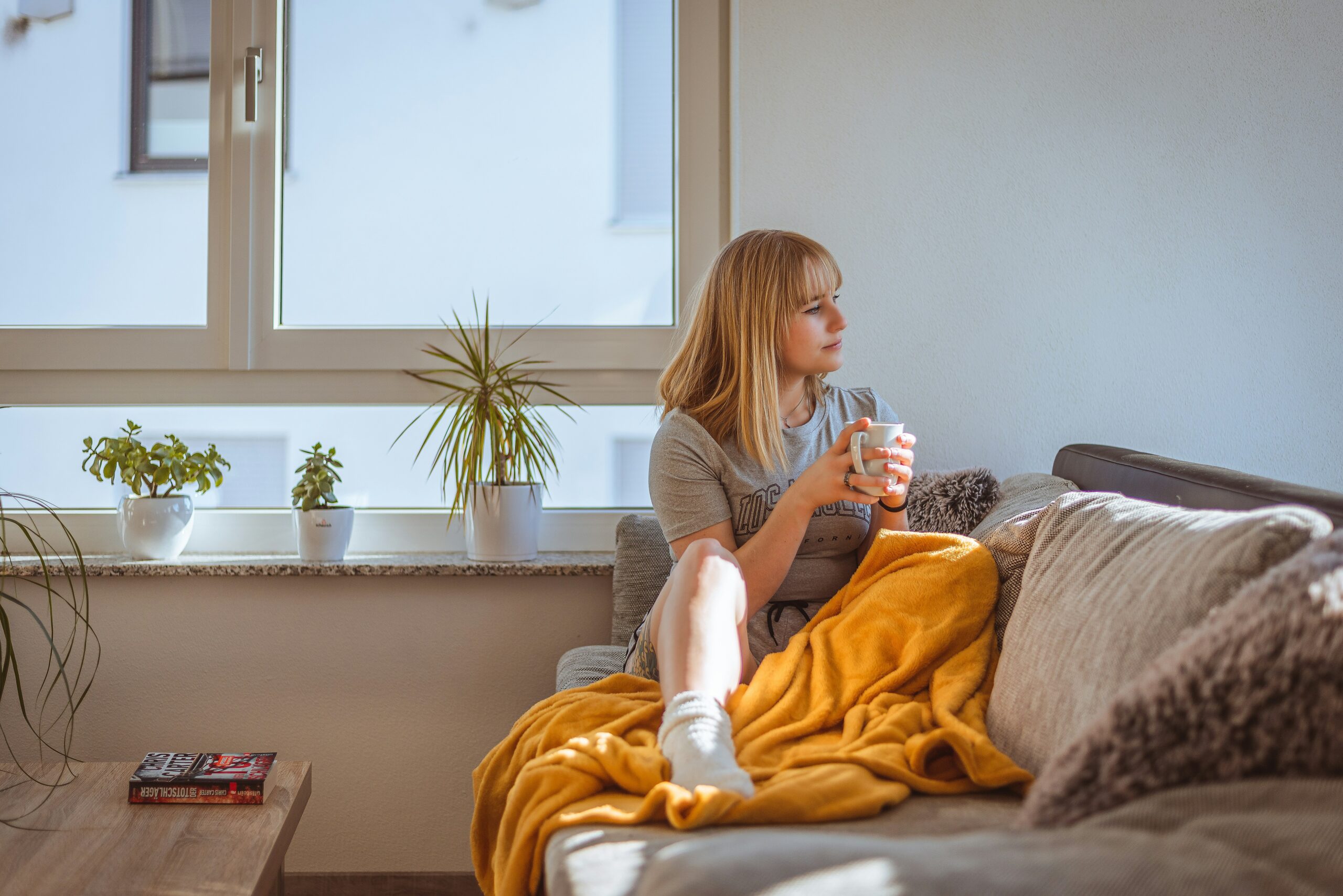 A woman sits under a yellow blanket on a couch holding a mug. A book sits on a table nearby.