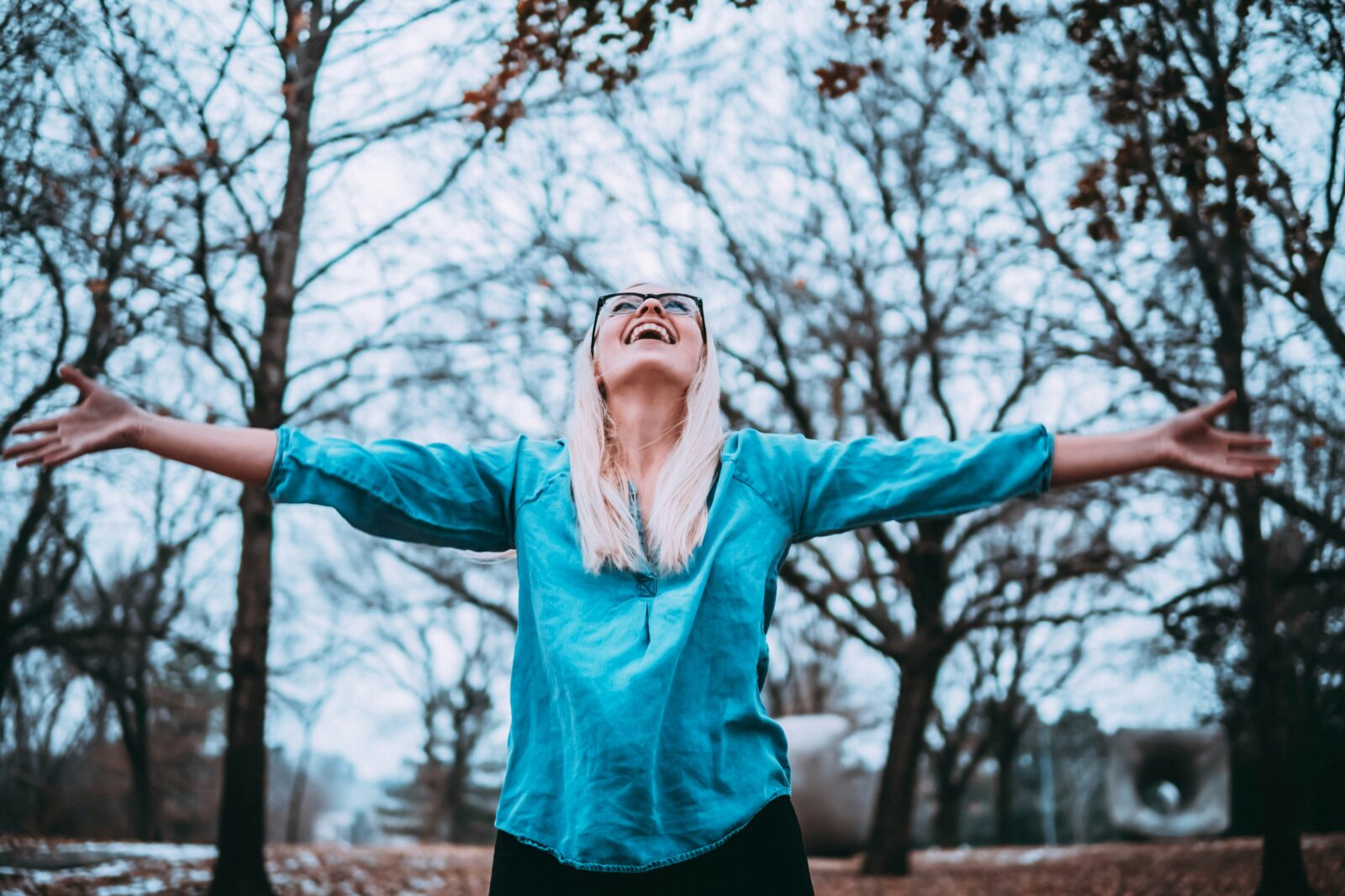 Woman in a teal shirt stands with arms outstretched amongst tall trees - breathing