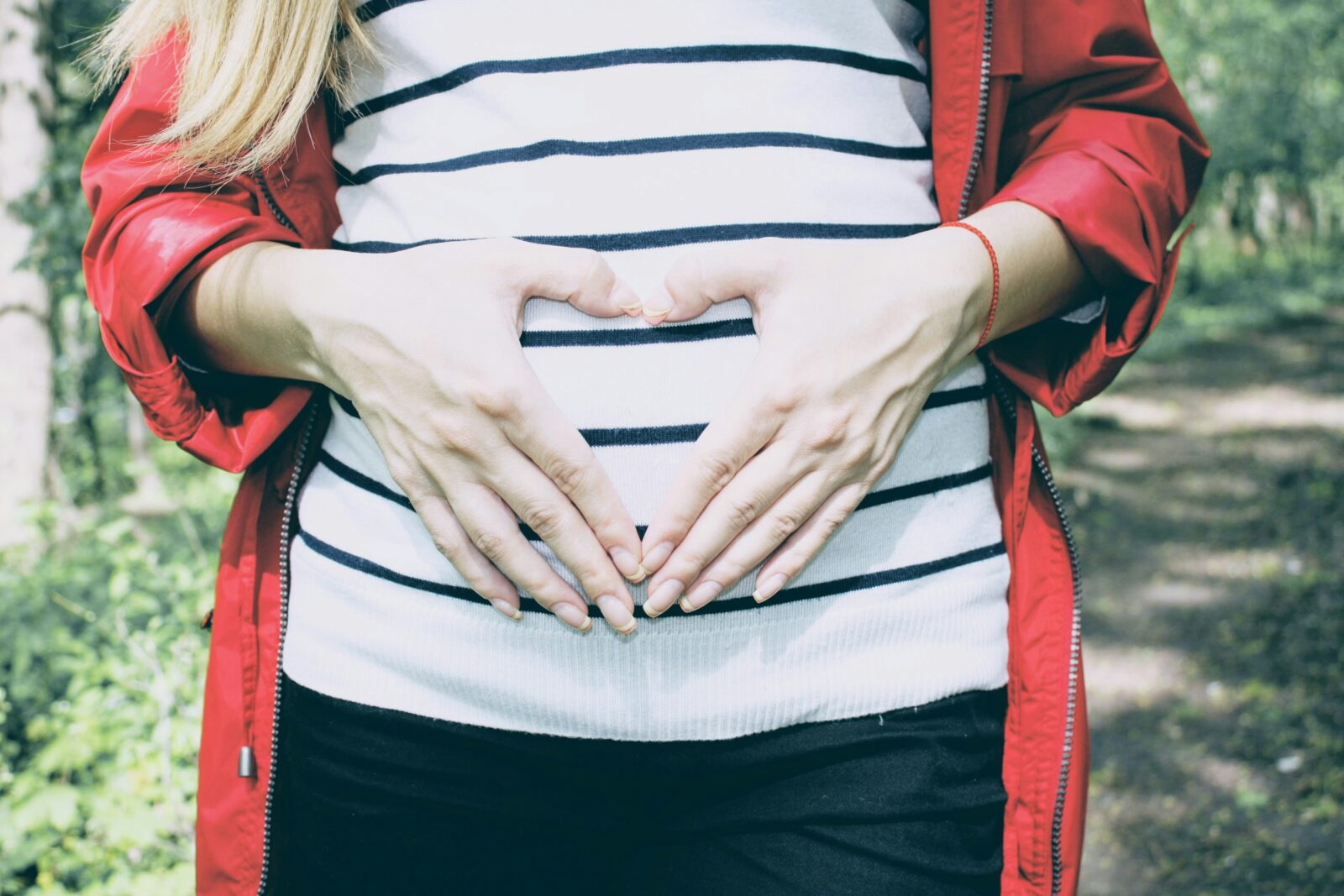 A woman in a striped top and red jacket holds her hands in a heart shape over her abdomen.